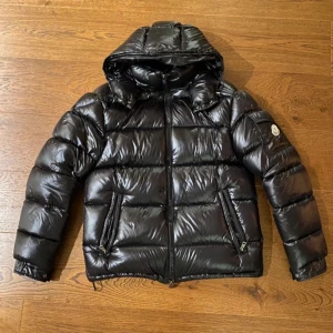 Svart dunjacka från Moncler - Säljer en snygg och varm svart dunjacka från Moncler. Jackan har en glansig finish och är perfekt för kalla vinterdagar. Den har en dragkedja framtill och en huva för extra skydd mot väder och vind. Märkesloggan syns tydligt på ärmen. Perfekt för dig som vill hålla stilen i vinter! 🖤