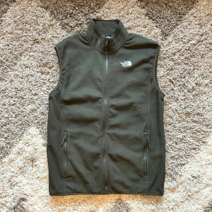 Grön fleeceväst, The North Face - Säljer en grön fleeceväst från The North Face i mycket bra skick. Västen har en dragkedja framtill och två praktiska fickor med dragkedjor. Perfekt för höst och vår när du vill hålla dig varm utan att bli för varm. Passar både för träning och vardagsbruk.