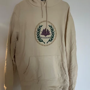 Beige hoodie från Woodbird - Säljer en snygg beige hoodie från Woodbird i storlek S. Den har en cool logga med en lila blomma och gröna blad på framsidan. Perfekt för en avslappnad stil och gjord av mjukt material för extra komfort. Passar perfekt till höst och vår! Tröjan har några mindre fläckar på framsidan som borde gå att få bort enkelt i tvätten