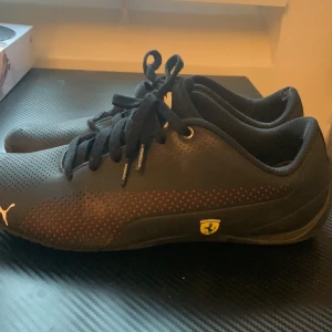 Svarta sneakers med Ferrari-logga - Snygga svarta sneakers från Puma med en cool Ferrari-logga på sidan. Skorna har snörning och små röda detaljer som ger en sportig look. Perfekta för dig som gillar både stil och komfort. De är i mycket bra skick och redo för nya äventyr!