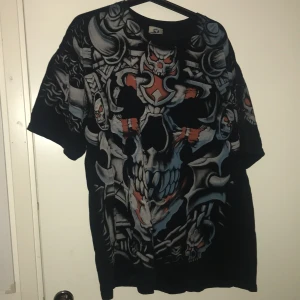 Svart t-shirt med dödskallemotiv från Liquid Blue - Cool svart t-shirt från Liquid Blue med ett stort, detaljerat dödskallemotiv i grått och rött. Perfekt för dig som gillar en edgy stil. T-shirten är kortärmad och gjord i bomull, vilket gör den bekväm att bära. Passar perfekt till både vardag och fest!