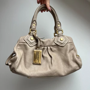 Marc by Marc Jacobs väska - Säljer en superfin beige handväska från Marc by Marc Jacobs i skinn. Väskan har snygga gulddetaljer och en praktisk dragkedja. Väskan är i fint skick 🤎 Mått bredd 30cm höjd 19cm, detta är den minsta modell