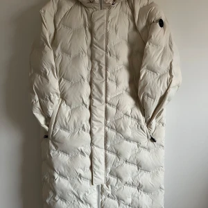 Vit lång dunjacka från Pelle P - Nypris: 2800kr. Säljer en supermysig vit lång dunjacka som är perfekt för kalla vinterdagar. Jackan har en stilren design med dragkedja och huva för extra värme. Den är i mycket bra skick och passar perfekt för både vardag och vinteräventyr. Håll dig varm och snygg i vinter! ❄️