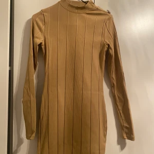 Beige ribbad klänning - Snygg beige klänning i ribbat material med långärmad design. Perfekt för höst och vinter med sin varma ton och stilrena look. Klänningen har en tight passform som framhäver figuren. Saknar bara två knappar uppe på halsen men de märks inte.