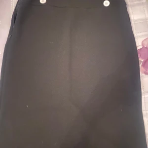 Svart kjol med knappar - Snygg svart kjol i mycket bra skick. Den har en klassisk design med två vita knappar framtill som ger en stilren look. Perfekt för både vardag och festliga tillfällen. Passar bra till en blus eller en enkel t-shirt.