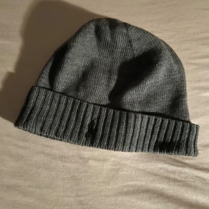 Grå Polo mössa - Säljer en enkel och stilren grå stickad Polo mössa. Perfekt för kyliga dagar med sin ribbade kant som håller den på plats. Mössan är i bra skick och passar de flesta huvudstorlekar. Ett måste för höst- och vintergarderoben!