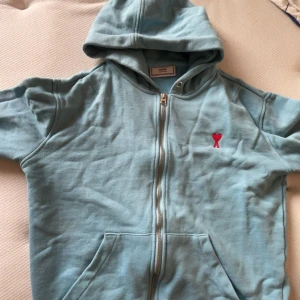 Ami Paris hoodie med dragkedja. - Säljer en ljusblå hoodie från Ami i mycket bra skick. Den har en dragkedja framtill och en liten röd logga på bröstet. Perfekt för vår och sommar, med korta ärmar och en skön passform. Huvan ger en avslappnad look. Perfekt för chill dagar eller en promenad i parken.