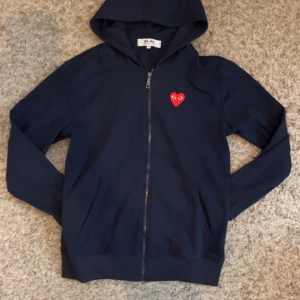 Svart hoodie från Comme des Garçons - Säljer en snygg svart hoodie från Comme des Garçons med det ikoniska röda hjärtat på bröstet. Den har en dragkedja framtill och är perfekt för både höst och vår. Superbekväm och stilren design som passar till allt. Perfekt för dig som vill ha något unikt och trendigt! 🖤❤️