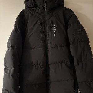Svart dunjacka från Sail Racing - Säljer en svart dunjacka från Sail Racing i mycket bra skick. Jackan har en stilren design med dragkedjor och en logga på bröstet. Perfekt för kalla vinterdagar med sin varma och skyddande stil. Den har en justerbar huva och flera praktiska fickor. Passar både till vardags och äventyr! Jackan är även helt ny då jag köpte i fel storlek. Nypriset är 4500kr, nu säljer jag denna för 3500kr. Kvitto när jag köpte den finns också 