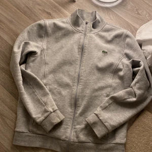 Lacoste Tröja - Fin lacoste tröja. Väldigt skön och fin för lågt pris. Nypris 1200 kr. Dragkedja till fickorna 