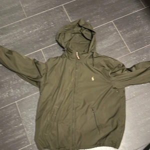 Olivgrön jacka från Polo Ralph Lauren - Säljer en snygg olivgrön jacka från Polo Ralph Lauren i storlek XL. Jackan har en broderad logga på bröstet och är tillverkad i polyester. Perfekt för höst och vår med sin praktiska huva och dragkedja framtill. Den är i mycket bra skick och redo för nya äventyr! 🍂Kom med bud. Den passar runt 185-195 cm