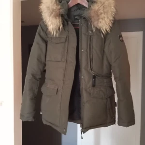 Olivgrön parkajacka med pälsdetaljer - Säljer en snygg olivgrön parkajacka från Hollies. Jackan har en mysig pälsfodrad huva och praktiska fickor framtill. Perfekt för kalla vinterdagar med sin långa ärm och varma material. Den har en dragkedja framtill och en stilren design som passar både till vardags och fest.