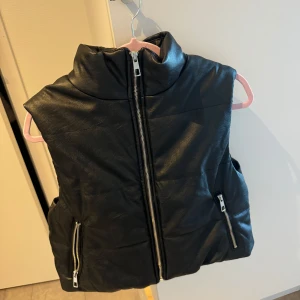 Svart skinnväst med dragkedja - Snygg svart skinnväst i mycket bra skick. Västen har en dragkedja framtill och två fickor med dragkedjor. Perfekt för höst och vår, och ger en cool look till vilken outfit som helst. Passar både till vardags och fest!