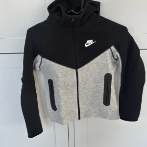 Svart och grå hoodie från Nike - Säljer en snygg och bekväm hoodie från Nike i svart och grått. Den har en dragkedja framtill och två praktiska fickor med dragkedjor. Perfekt för höst och vår, eller när du bara vill ha något mysigt att ha på dig. Passar bra till både träning och vardag. Storlek M barn