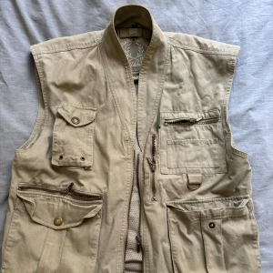 Beige Chevalier Jägarväst - Vintage beige jägarväst från Chevalier med ”double access” ficka i ryggen Ärvt från äldre familjemedlem som är f.d jägare, troligt flitigt använd, utan synliga defekter Säljer då den ej faller mig i smaken
