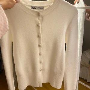 Vit kofta från ZARA - Superfin vit kofta från ZARA i mycket bra skick. Den är långärmad och har snygga glittriga knappar framtill. Perfekt för både höst och vinter, och den är gjord i ett mjukt material som känns lyxigt. Passar perfekt till både vardag och fest! Skit snygg men tyvärr lite liten för mig över brösten. Använd kanske 2 gånger men är i superfint skick! Skulle säga att den är mer som en S.