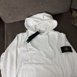 Vit hoodie från Stone Island - Säljer en snygg vit hoodie från Stone Island i nytt skick. Den har en klassisk design med en känguruficka och en justerbar huva. Perfekt för både vardag och chill. Märkesloggan sitter på ärmen för en stilren look. Passar perfekt till jeans eller joggers!