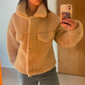 Beige teddyjacka från Gina - Säljer en supermysig beige teddyjacka från Gina i mycket bra skick. Den har en normal passform och långa ärmar, perfekt för höst och vinter. Jackan har knappar framtill och är gjord av ett mjukt och varmt material. Passar S/M