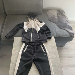 Svarta mjukisbyxor från Boss - Säljer ett par svarta mjukisbyxor från Boss med gråa detaljer längs sidorna. Byxorna har en elastisk midja med snörning och två fickor fram. Perfekta för en avslappnad stil eller träning. De är i mycket bra skick och superbekväma! Nästan aldrig använda:)