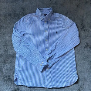 Blå och vit randig skjorta från Polo Ralph Lauren - Säljer en snygg blå och vit randig skjorta från Polo Ralph Lauren i slim fit. Skjortan har långa ärmar och knappar framtill. Pris kan diskuteras vid snabb äffär. Storlek 164 men passar som xs.
