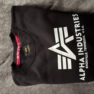 Svart sweatshirt från Alpha Industries - Säljer en svart sweatshirt från Alpha Industries med deras ikoniska logga i vitt på framsidan. Tröjan är i mycket bra skick och perfekt för både vardag och chill. Den har en klassisk rund hals och är superbekväm. Passar perfekt till jeans eller joggers!