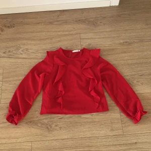 Röd blus med volangdetaljer - Snygg röd blus i storlek 146 med volangdetaljer framtill. Perfekt för både vardag och festliga tillfällen. Blusen har långa ärmar och en bekväm passform. Den är gjord i ett mjukt material som känns skönt mot huden.