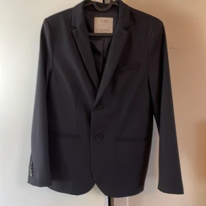 Svart kavaj från Zara Boys - Snygg svart kavaj från Zara Boys i storlek 152, perfekt för festliga tillfällen. Kavajen har en klassisk design med två knappar fram och fickor på sidorna. Tillverkad i Bulgarien, den är i nytt skick och redo att bäras till nästa stora event!