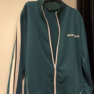 Palm Angels kofta  - Säljer en snygg grön track jacket från Palm Angels. Den har en vit dragkedja och vita ränder längs ärmarna. Perfekt för en sportig look eller casual vardagsstil. Jackan är i bra skick och redo för nya äventyr!