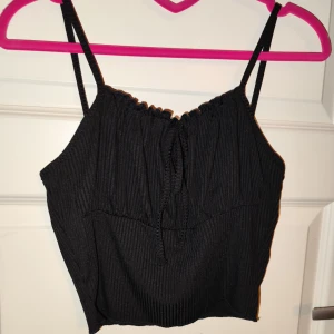 Svart ribbad topp - Säljer en snygg svart ribbad topp med justerbara axelband och knytning framtill. Perfekt för sommardagar eller en utekväll. Toppen är i mycket bra skick och passar till både jeans och kjol. Ett måste i garderoben!
