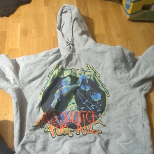 Grå Metallica hoodie - Säljer en grå Metallica hoodie med tryck från albumet '...And Justice for All'. Den har en stor färgglad grafik på framsidan och text på baksidan. Perfekt för fans av bandet! Hoodien är i bra skick och har en skön passform med långa ärmar och en klassisk huva.