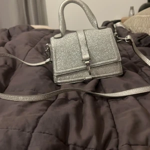 Glittrig silverväska från H&M - Säljer en superfin glittrig silverväska från H&M. Perfekt för fest eller nyår! Den har en praktisk axelrem och ett snyggt spänne framtill. Väskan är i mycket bra skick och redo att användas för att ge din outfit det där lilla extra. ✨