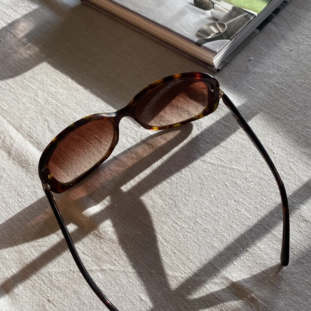 Säljer ett par snygga solglasögon från Prada. De har en klassisk brun färg med ett stilrent mönster som påminner om sköldpaddsskal. Glasögonen har en rektangulär form och är perfekta för soliga dagar. De är i bra skick och har en elegant design med Pradas logga på sidan. Perfekta för att ge din outfit en lyxig touch!. Asusteet.