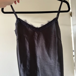 Svart siden linne med spetsdetaljer - Säljer en elegant svart linne i siden med fina spetsdetaljer vid halsringningen och nederkanten. Klänningen har smala axelband och en mjuk, glansig yta som ger en lyxig känsla. Perfekt för en festlig kväll eller en speciell tillställning. 🖤