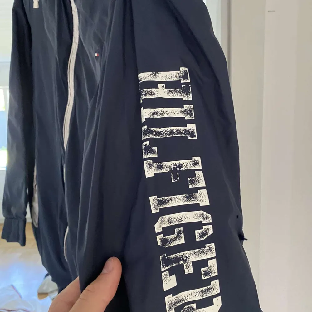 Säljer en snygg mörkblå vindjacka från Tommy Hilfiger. Jackan har en dragkedja framtill och vita detaljer vid fickorna. På ärmen finns det ett stort tryck med 'HILFIGER'. Perfekt för vår och höst!. Jackor.