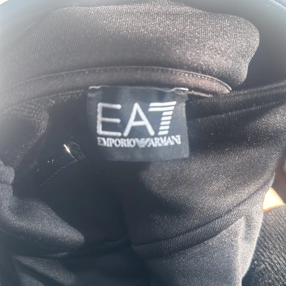 Säljer en snygg svart jacka från Emporio Armani med coola guldfärgade detaljer. Jackan har en dragkedja framtill och EA7-loggan på bröstet. Perfekt för en stilren look!. Hoodies.