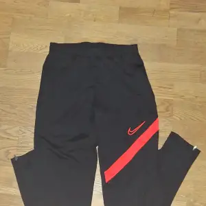 Säljer ett par svarta träningsbyxor från Nike i storlek S. Byxorna är tillverkade i Dri-FIT-material som håller dig torr och bekväm under träningen. De har en röd Nike-logga och en röd rand längs sidan. Perfekta för gymmet! Pris: 30kr