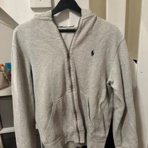 Grå hoodie från Polo Ralph Lauren - Säljer en snygg och bekväm grå hoodie från Polo Ralph Lauren. Den har en dragkedja framtill och en klassisk logga på bröstet. Perfekt för en avslappnad stil och passar till  alla tillfällen. Den är i bra skick och redo för en ny ägare! Passar även S
