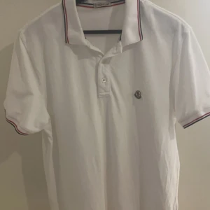 Moncler Piké - Säljer en stilren vit pikétröja från Moncler. Den har en klassisk krage med röd och blå randdetalj samt en liten logga på bröstet. Perfekt för en casual look eller en dag på golfbanan. Tröjan är i bra skick och redo för nya äventyr!