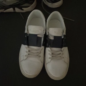 Valentino Garavani sneakers - Säljer ett par snygga Valentino Garavani sneakers i vitt med svarta detaljer. Skorna har en stilren design med snörning och är i skinn. De är i välanvänt skick men fortfarande riktigt coola och perfekta för både vardag och fest. 