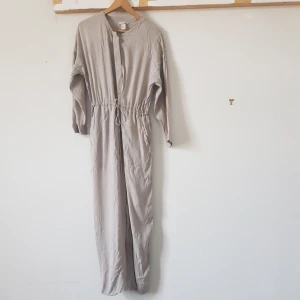 Beige lång jumpsuit - Säljer en stilren och bekväm gråbeige jumpsuit i storlek XS. Den har långa ärmar och en dragsko i midjan för en justerbar passform. Perfekt för en avslappnad dag eller en stilfull kväll.