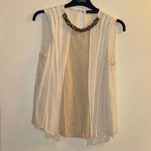 Vit ärmlös topp från Zara - Säljer en elegant vit ärmlös topp från Zara. Den har en vacker pärldetalj vid halsringningen som ger en lyxig touch. Toppen är i ett lätt och flödande material, perfekt för sommarens fester eller en stilren vardagslook.
