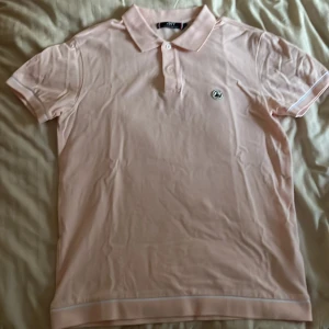 Ljusrosa pikétröja från JOTT - Säljer en snygg ljusrosa pikétröja från JOTT. Den har en klassisk krage med knappar och ett litet märke på bröstet. Perfekt för en avslappnad men stilren look. Passar bra till både vardag och fest!