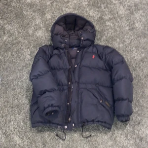 Mörkblå dunjacka från Ralph Lauren - Säljer en snygg och varm mörkblå dunjacka från Ralph Lauren. Jackan har en klassisk pufferstil med en huva och dragkedja framtill. Den är dekorerad med det ikoniska röda Polo-logotypen på bröstet. Perfekt för kalla vinterdagar!