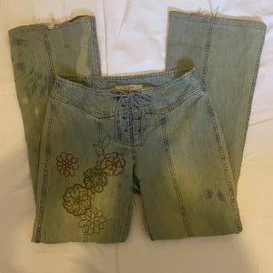 Coola lågmidjade jeans med snörning i midjan - Ett par lågmidjade bootcut jeans som har snörning i midjan. De har broderade blommor på ena benet och är fläckigt bleka. Tyvvär har de lite slitningar längst ner på benen, om du har några frågor eller vill ha flera bilder är det bara att fråga💕