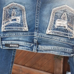 Blå bootcut jeans med paljettdetaljer - Säljer ett par snygga blå bootcut jeans från VICOSS. De har coola paljettdetaljer och slitningar på bakfickorna som ger en unik look. Jeansen har en klassisk femficksdesign och är perfekta för en avslappnad stil. Liten fläck på byxbenet .M 40cm IL 78