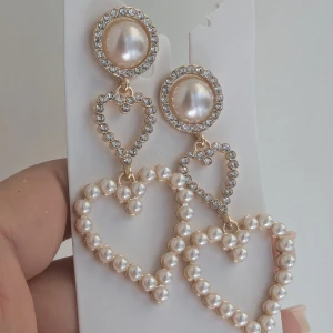 Pärlörhängen med hjärtan och strass - Superfina örhängen med stora pärlor och hjärtformade detaljer. De har en rund pärla i mitten omgiven av glittrande strass, och hängande hjärtan dekorerade med små pärlor.NYA