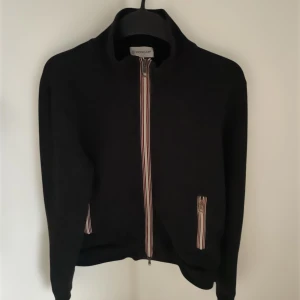 Svart Moncler Maglia zip (äkta) - Snygg svart cardigan från Moncler med dragkedja och ribbade detaljer. Jackan har en klassisk design med Moncler-logga på ärmen och praktiska fickor framtill. Perfekt för en stilren look.