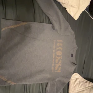 Grå tröja från Boss - Säljer en stilren grå tröja från Boss med ett glittrigt logotyptryck på bröstet. Tröjan har långa ärmar och en klassisk passform. Perfekt för en avslappnad men ändå snygg look. Köpt på season i asecs.