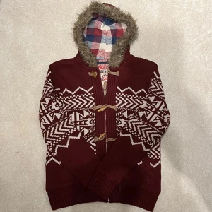 Fodrad Cardigan Jacka med päls - Säljer en vinröd stickad jacka med ett snyggt geometriskt mönster i vitt. Jackan har en huva med fuskpäls och ett rutigt innerfoder. Den stängs med träknappar och har ribbade muddar vid ärmslut och nederkant. Vid minsta fråga eller fundering hör av er🤝