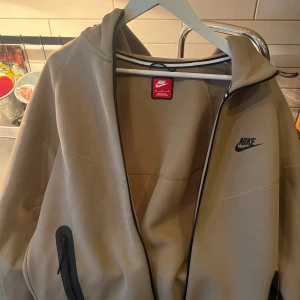 Zip hoodie jacka från Nike - Säljer en snygg beige zip hoodie från Nike med dragkedja och svart logga på bröstet. Zip hoodien har långa ärmar och en stilren design, perfekt för vardagsbruk. Den är tillverkad i ett syntetmaterial som ger en modern look.